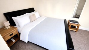 A neatly made bed with white and beige bedding, a wooden nightstand with a lamp, and a small wooden table with a book and a bottle. - City Stay Worcester - free parking (Worcester)
