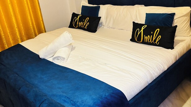 Een net opgemaakt bed met een donkerblauwe hoofdbord, witte beddengoed en twee kussens met de tekst 'Smile'.
