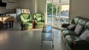 A living room with a green leather sofa, a glass coffee table, and a dining table with chairs. There is a kitchen area with a refrigerator and a window.