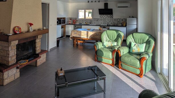 A spacious living room with a fireplace, green leather armchairs, a glass coffee table, and a kitchen in the background.