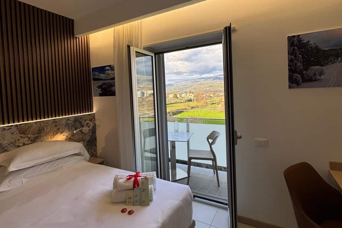 A hotel room with a bed, a desk, a chair, and a balcony with a view of a cityscape.