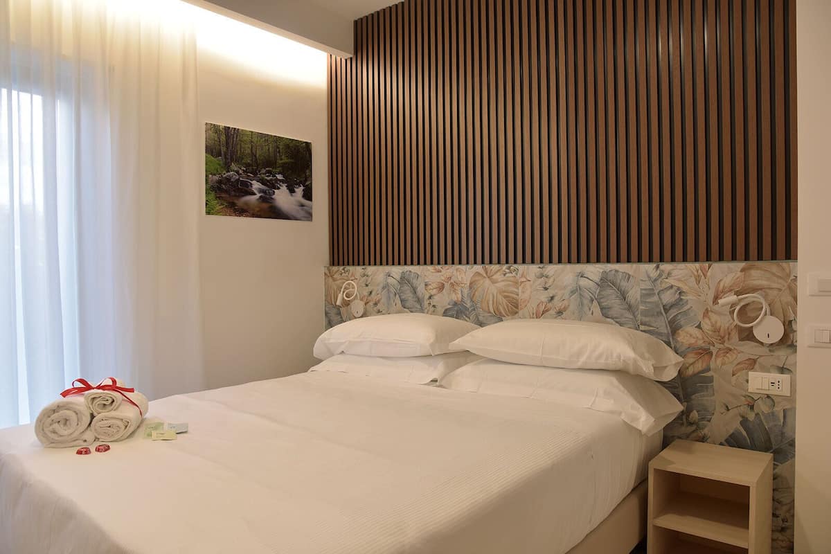 A neatly made bed with white linens, a wooden headboard with vertical slats, a floral-patterned wall hanging above the bed, and a small wooden nightstand beside the bed.