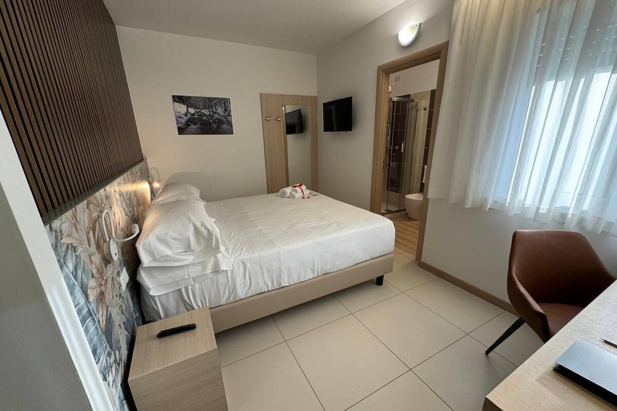 A hotel room with a bed, a desk with a laptop, a chair, a TV, and a shower area.