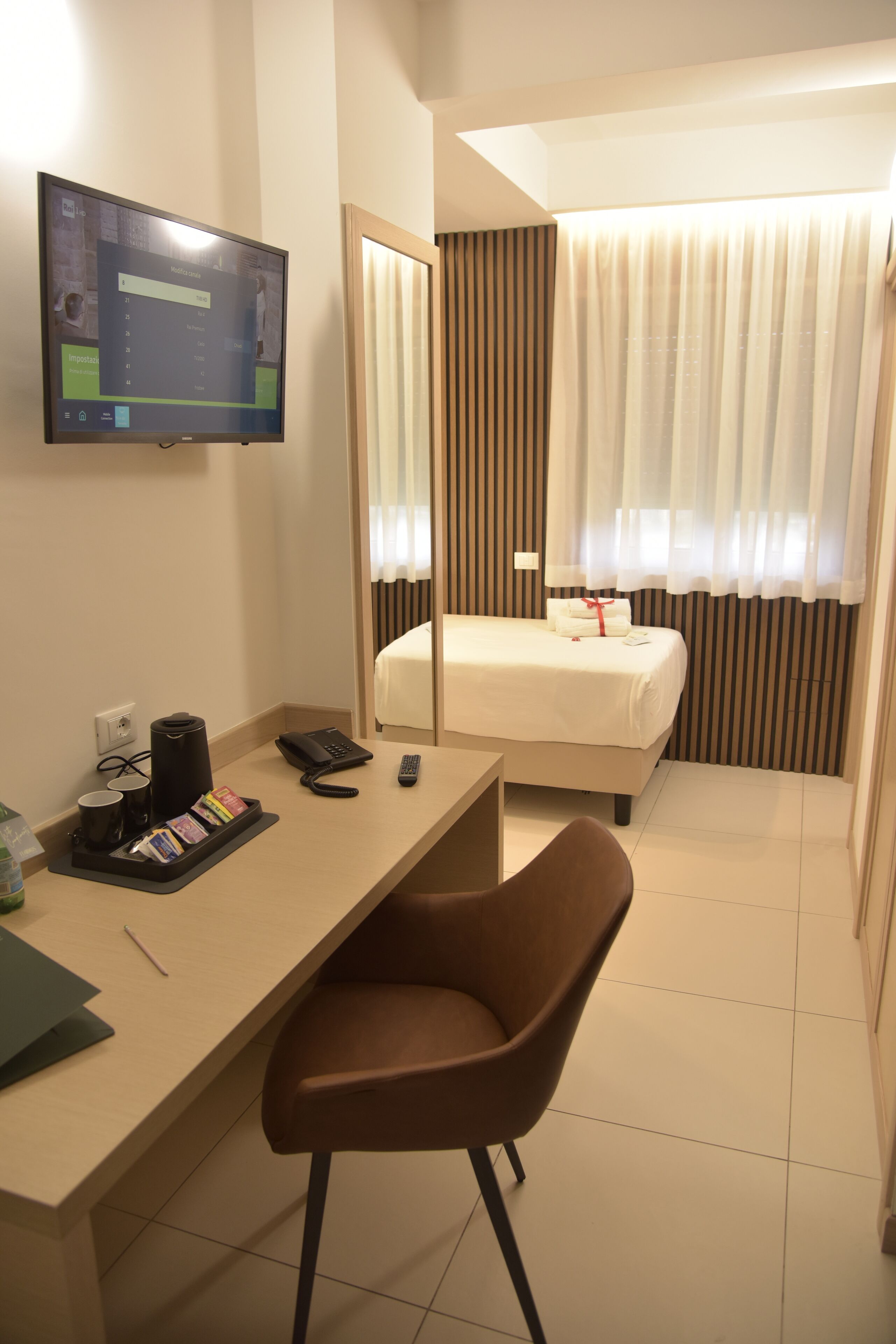 A hotel room with a bed, a desk with a chair, a TV, and a window with curtains.