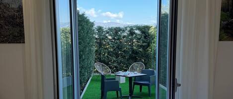 Quarto de hotel com cama, escrivaninha e vista para área externa com mesa e cadeiras.