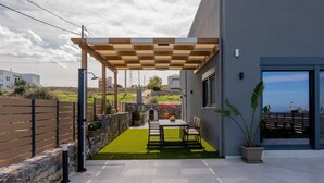 A modern outdoor patio with a pergola, a stone wall, and a view of the sea. - Villa Grand Blue (Hersonissos)
