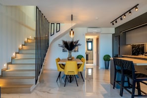 A modern kitchen with a central island, a dining area with yellow chairs, and a staircase with built-in lighting. - Villa Grand Blue (Hersonissos)