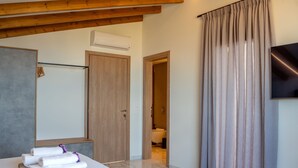 A hotel room with a wooden ceiling, a flat-screen TV, a bathroom visible through an open door, and a table with towels. - Villa Grand Blue (Hersonissos)