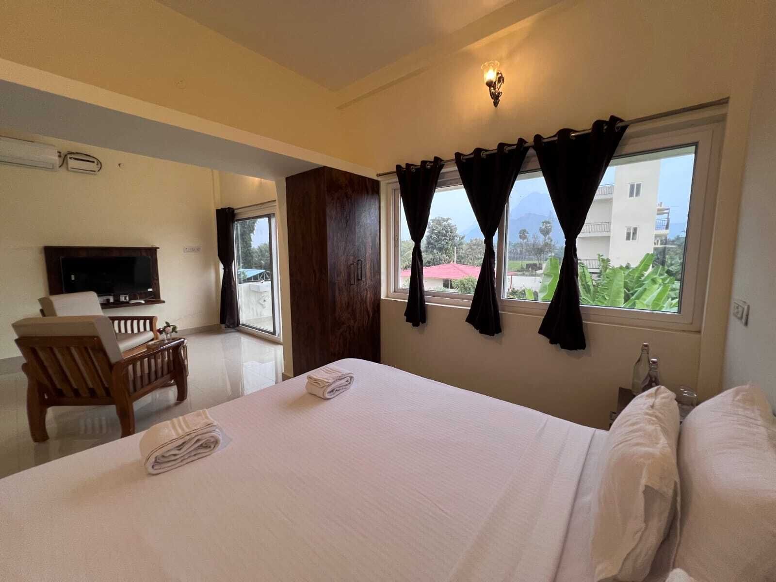 A hotel room with a bed, a TV, a chair, and a window with curtains.