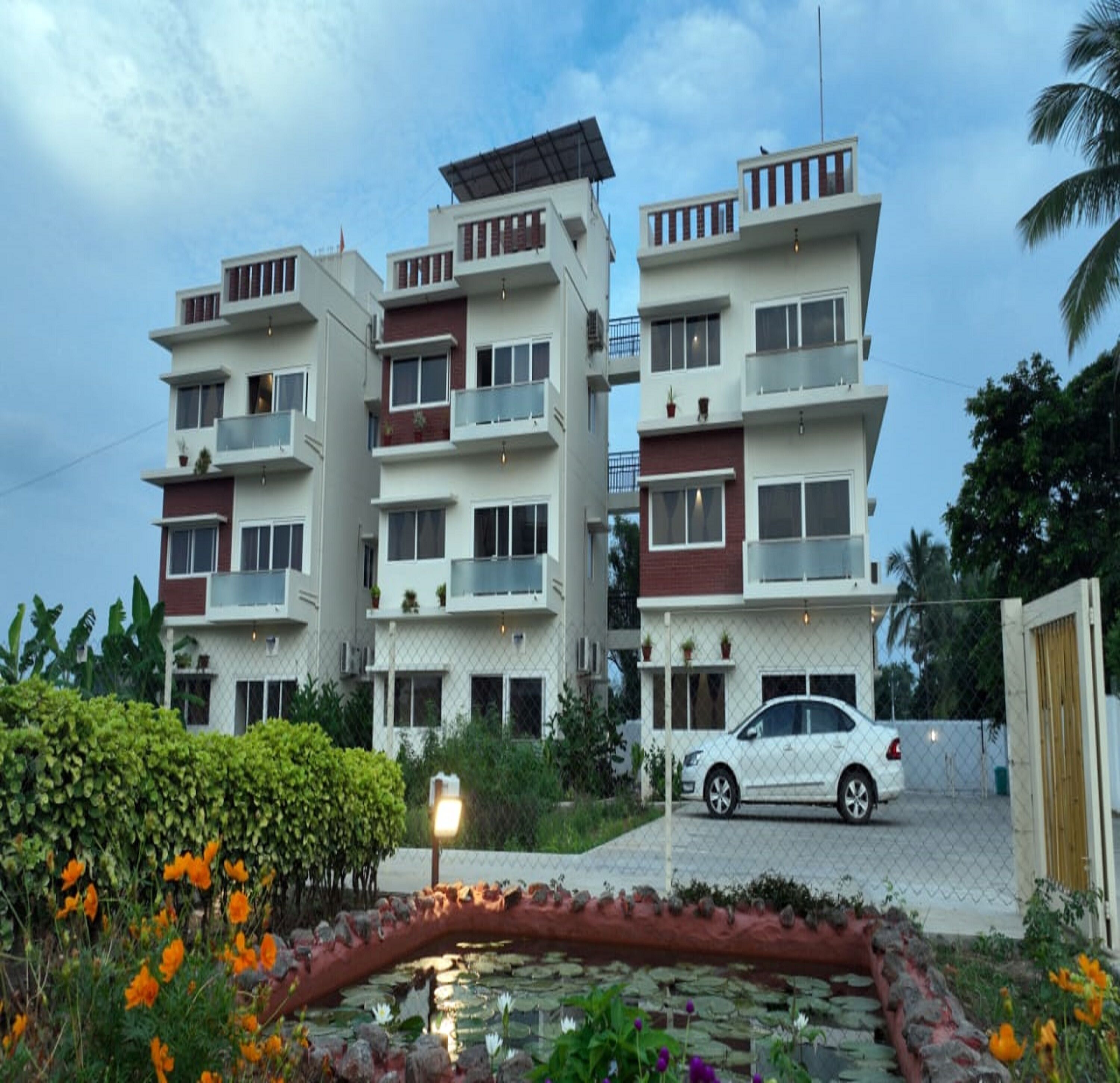 A multi-story residential building with balconies, a car parked in front, and a garden with flowers and a pond.