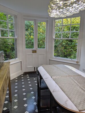 Une pièce avec une longue table, deux fenêtres donnant sur un paysage verdoyant, un lustre et un sol à motifs.