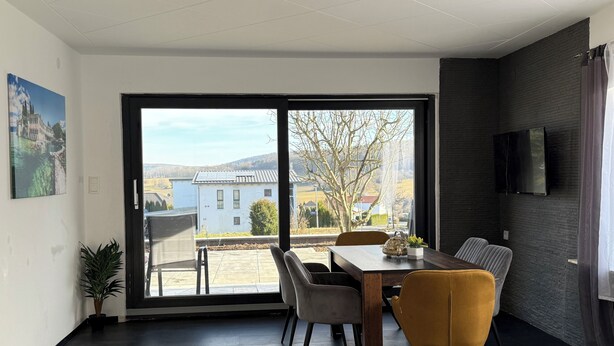 Un comedor moderno con una mesa grande y sillas, un televisor montado en la pared y vistas a una casa y un árbol a través del ventanal.