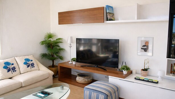A modern living room with a flat-screen TV, sofa, ottoman, glass coffee table, and decorative plants. - At469 in Oliva With 2 Bedrooms and 2 Bathrooms (Oliva)