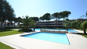 Una piscina con solárium de baldosas, rodeada de árboles y un edificio con balcones.