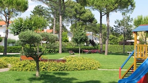 A playground with a slide and a bench in a park with trees and a building in the background. - Two-level flat with garden and pool (Bibione)