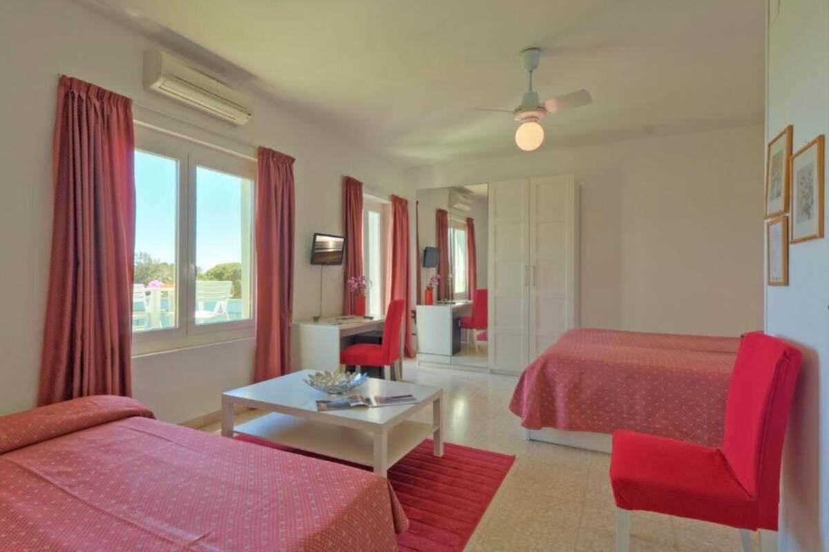 A room with two beds, a desk, a chair, a television, and a ceiling fan.