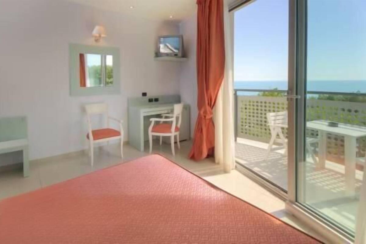 A bedroom with a bed, a desk, a chair, a TV, and a balcony with a view.