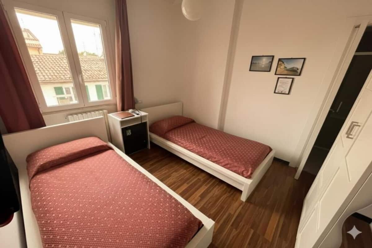 A bedroom with two beds, a nightstand, a radiator, a window with curtains, and a door.