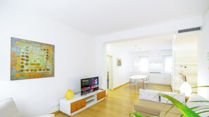 Uma sala de estar moderna com televisão de tela plana sobre um console branco, sofá bege e área de jantar com mesa e cadeiras brancas.