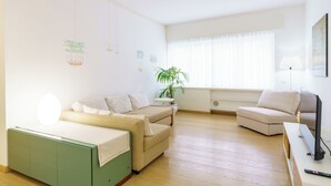 Uma sala de estar moderna com um sofá verde, poltronas bege, uma televisão e uma janela grande com cortinas leves.
