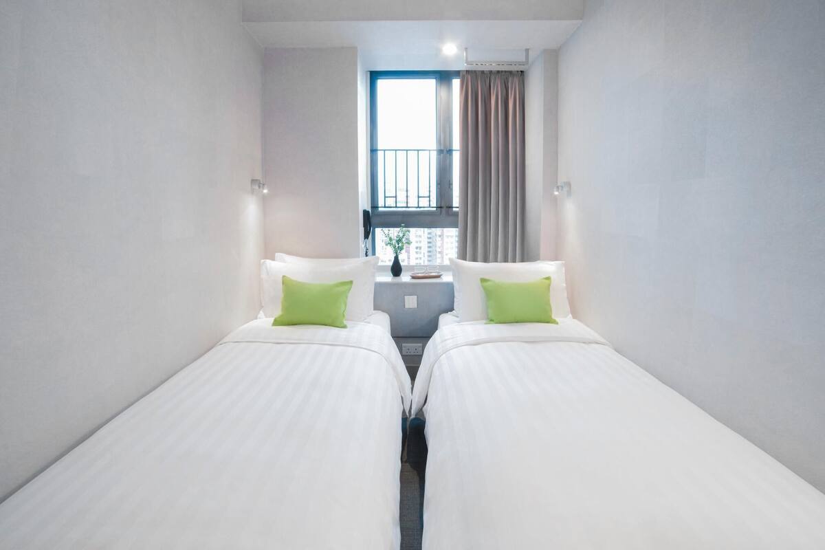Habitació d'hotel amb dos llits individuals, una taula petita amb un ram i gots, una finestra amb cortines, i vista a edificis a l'exterior.