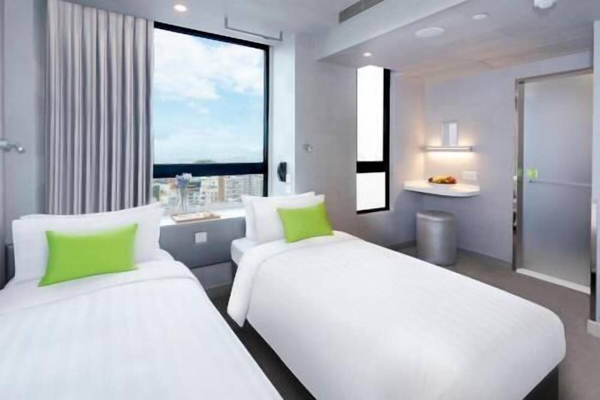 Una habitació d'hotel amb dos llits, una finestra que dona a una vista de la ciutat, un petit escriptori amb una làmpada i una porta.