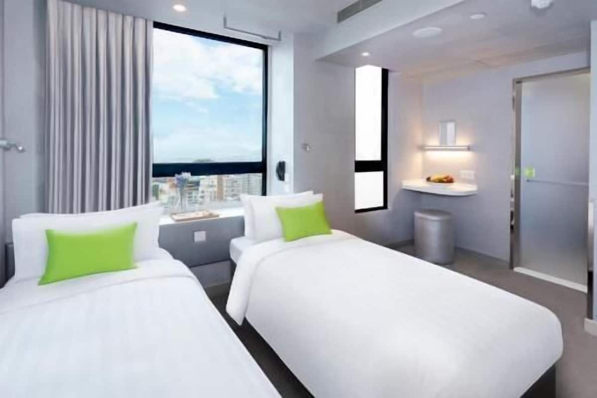 Ein Hotelzimmer mit zwei Betten, einem Fenster mit Blick auf die Stadt, einem kleinen Schreibtisch mit Lampe und einer Tür.