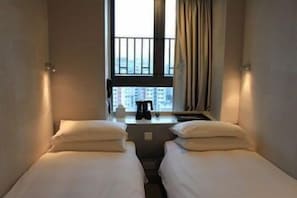A hotel room with two single beds, a small table with a lamp, and a window with curtains.