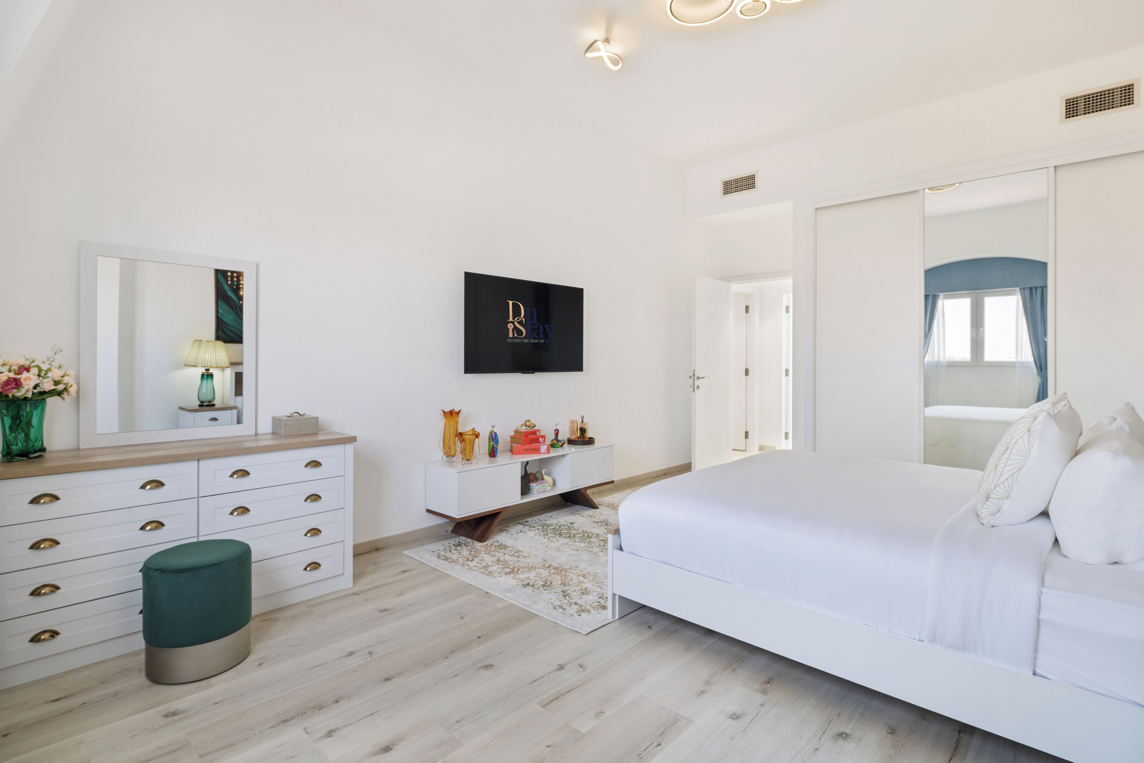 Ein modernes Schlafzimmer mit einem großen Bett, einer Kommode mit Schubladen, einem Spiegel und einem Flachbildfernseher an der Wand.