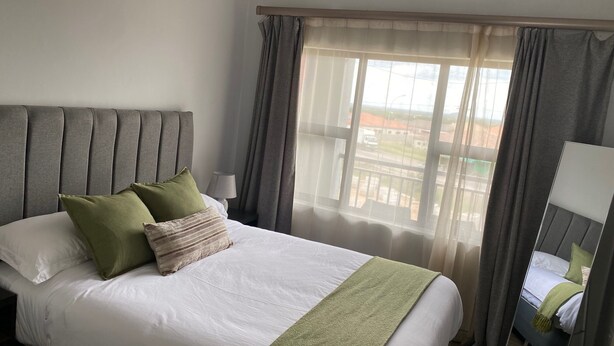 Habitación de hotel con una cama grande, una mesita de noche, una lámpara y vistas al exterior a través de cortinas traslúcidas.