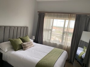Et hotelværelse med en stor seng, et natbord, en lampe og udsigt ud gennem lette gardiner.