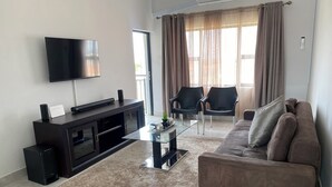 Una sala de estar moderna con un televisor de pantalla plana, un mueble multimedia oscuro, una mesa de centro de vidrio, un sofá marrón y dos sillas negras.