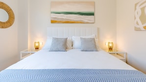 A bedroom with a large bed, two bedside tables with lamps, a wall-mounted painting, and a round mirror. - Bali in Rincón | Elegant 2BR Apartment with Terrace & Ocean Views (Rincón de la Victoria)