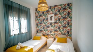 Een kamer met twee eenpersoonsbedden, een groot raam met blauwe gordijnen, tropische behang en een hanglamp.