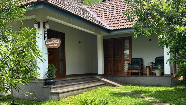 A house with a tiled roof, a porch with steps, a sign reading 'Blue Woods Home Stay,' and a small garden area. - Bluewoods Homestay – Serene Retreat Near Kochi City (Ernakulam)