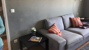 A living room with a grey sofa, two pillows, a magazine, a vase with flowers, and a framed picture on the wall. - Holiday Home 'Sunshine Home - 300m To The Beach' with Sea View, Private Terrace and Wi-Fi (Leça da Palmeira)