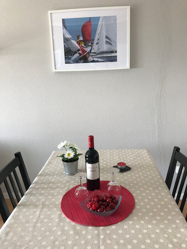 Une table à manger recouverte d’une nappe à pois, une bouteille de vin, un bol de cerises, une plante en pot et un tableau encadré accroché au mur.