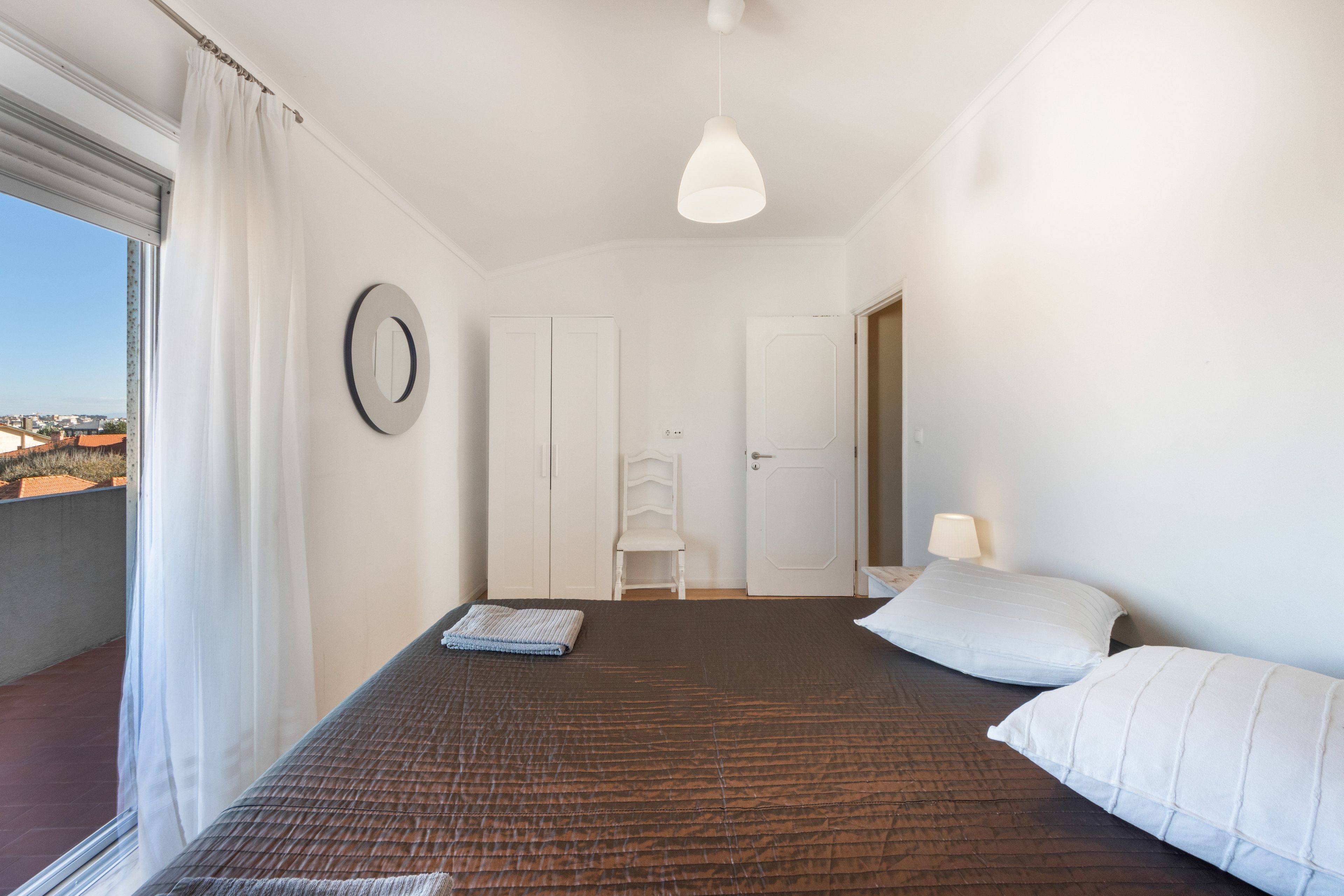 A bedroom with a bed, a bedside lamp, a window with curtains, a door, and a view of the city.