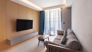 A modern living room with a flat-screen TV mounted on a wooden wall, a grey sofa, a small wooden coffee table, and sheer curtains on a large window. - Beachfront 5122 Building C Modern 2BR Suite (Choeng Thale)