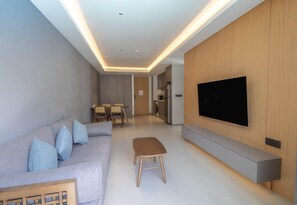 A modern living room with a sofa, a coffee table, a TV mounted on the wall, and a kitchen area in the background. - Beachfront 5122 Building C Modern 2BR Suite (Choeng Thale)