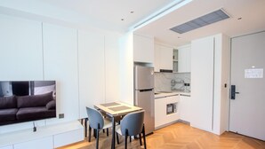 Une cuisine moderne avec des armoires blanches, un four encastré et un réfrigérateur. On y trouve une table à manger avec des chaises, ainsi qu’un téléviseur  à écran plat fixé au mur.