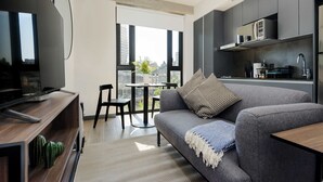 Un salon moderne avec un canapé gris, une table basse et un téléviseur posé sur un meuble en bois. On y trouve également un coin cuisine équipé d’un four à micro-ondes et d’un évier.
