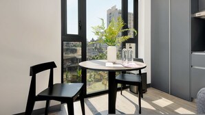 Une pièce moderne avec une table ronde, deux chaises noires, un vase blanc orné d’une plante, et une vue sur le paysage urbain à travers de grandes fenêtres.