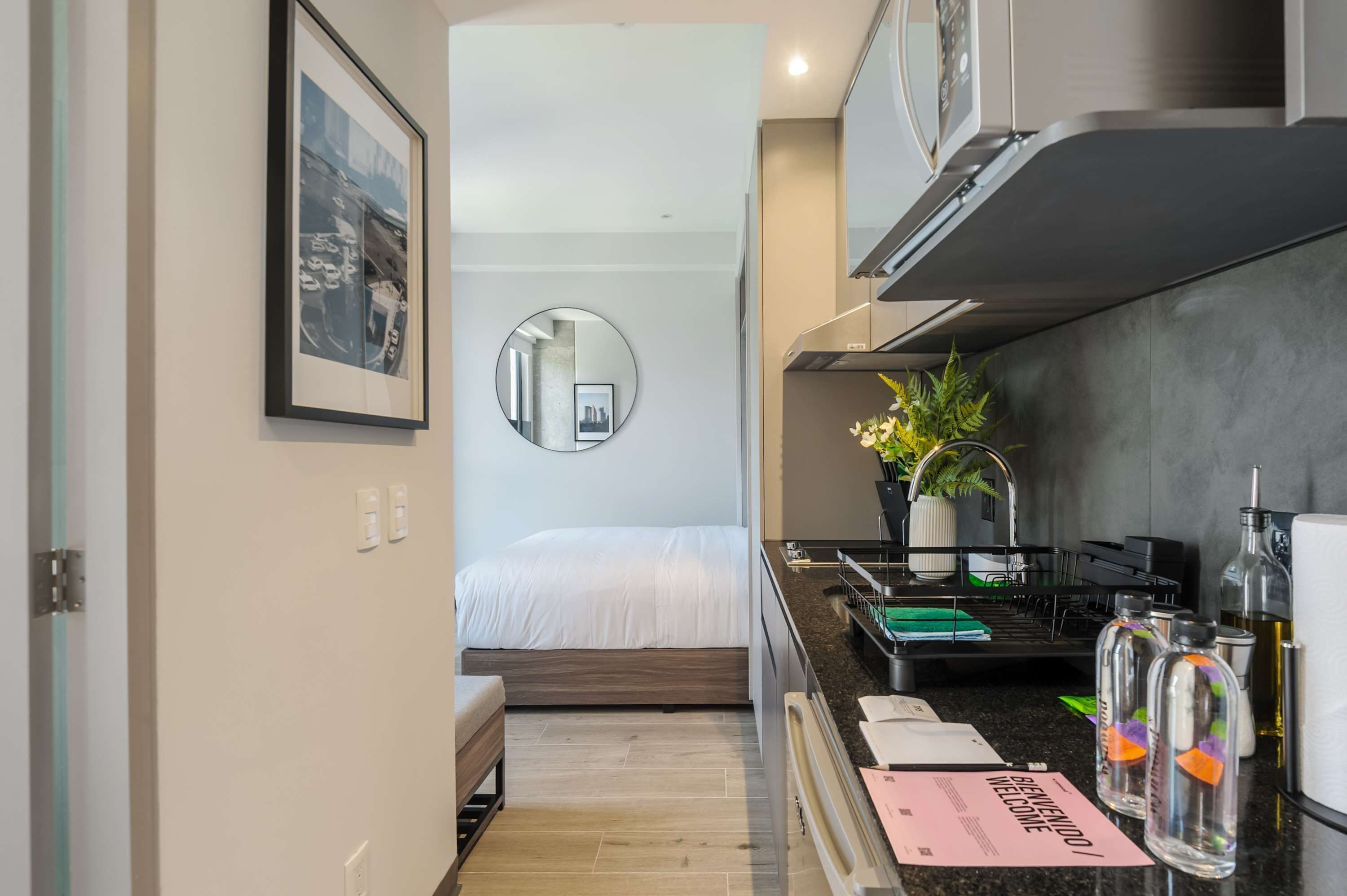 A compact hotel room with a bed, a small kitchenette, a round mirror, and a welcome card on the counter.