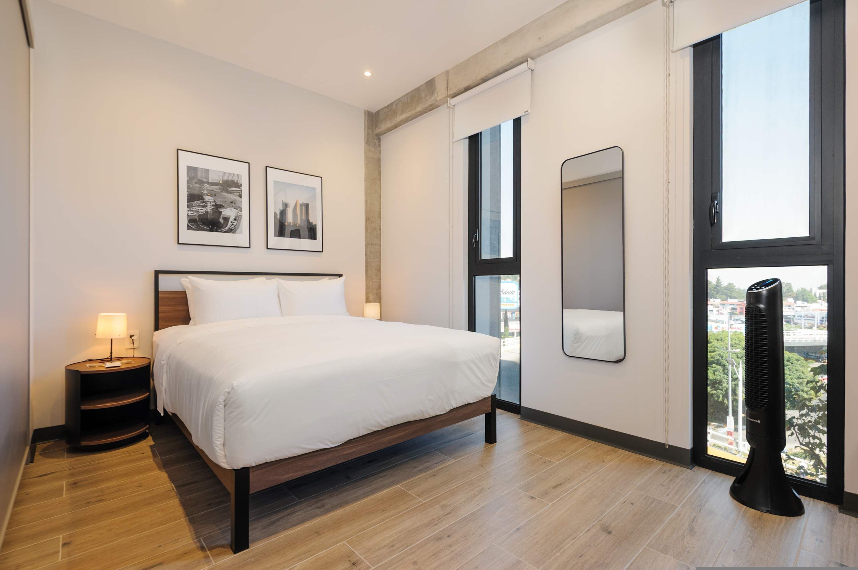 A modern hotel room with a large bed, a bedside table with a lamp, two framed pictures on the wall, a large window with a view, and a standing fan.