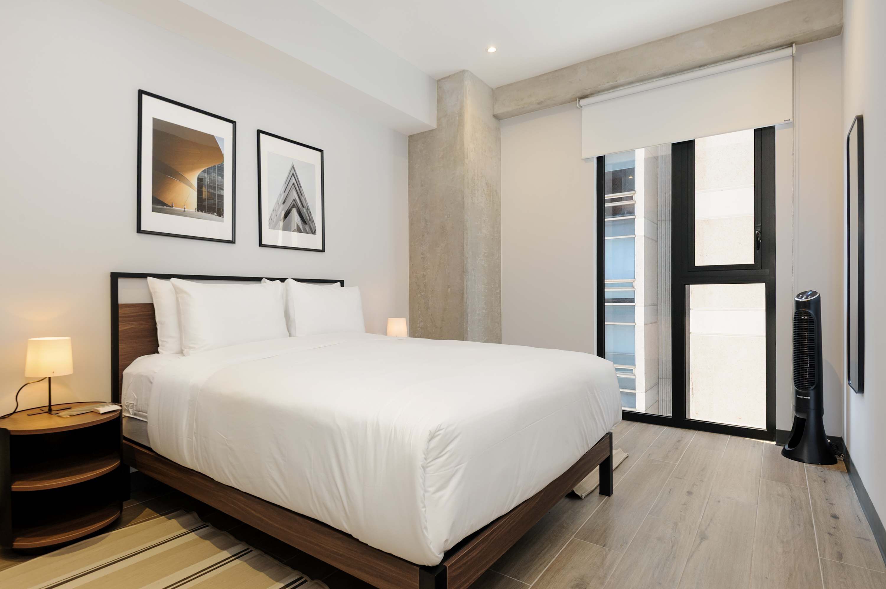 A modern hotel room with a large bed, a nightstand with a lamp, a black standing heater, and a large window with a view of the city.