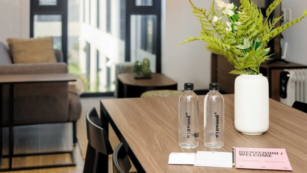 A wooden table with two clear water bottles, a white vase with green foliage, and a welcome card.