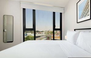 A hotel room with a large bed, a view of the cityscape through floor-to-ceiling windows, a framed picture on the wall, and a side mirror. - w Luxurious 2BR in the Heart of Interlomas (Huixquilucan)