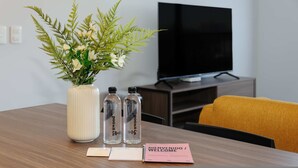 A room with a wooden table, a potted plant, two water bottles, a pink welcome card, and a TV on a stand. - w Charming 2BR in the Heart of Interlomas (Huixquilucan)