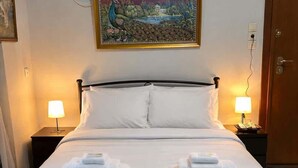 A neatly made bed with white linens and two folded towels, a painting above the bed, and a door with an exit sign. - Zografou Cozy Apartment in the heart of Athens! (Zografou)